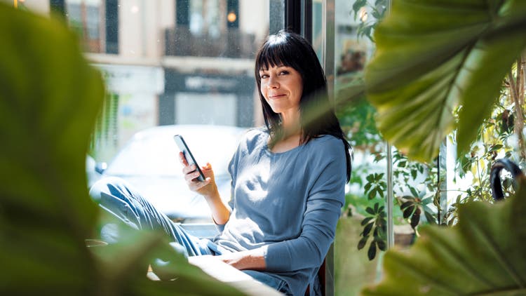 A woman sits by a window surrounded by plants, holding a smartphone