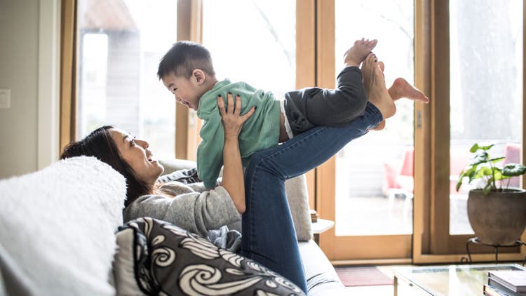 A woman lying on sofa lifts a smiling young boy above her with her legs